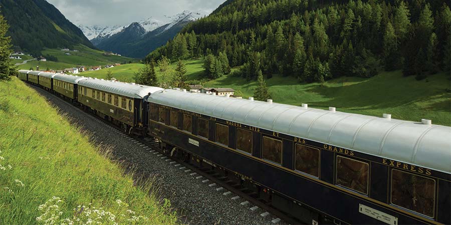 The Venice Simplon Orient Express travelling through a rural area with snow-capped mountains and trees in the background. 